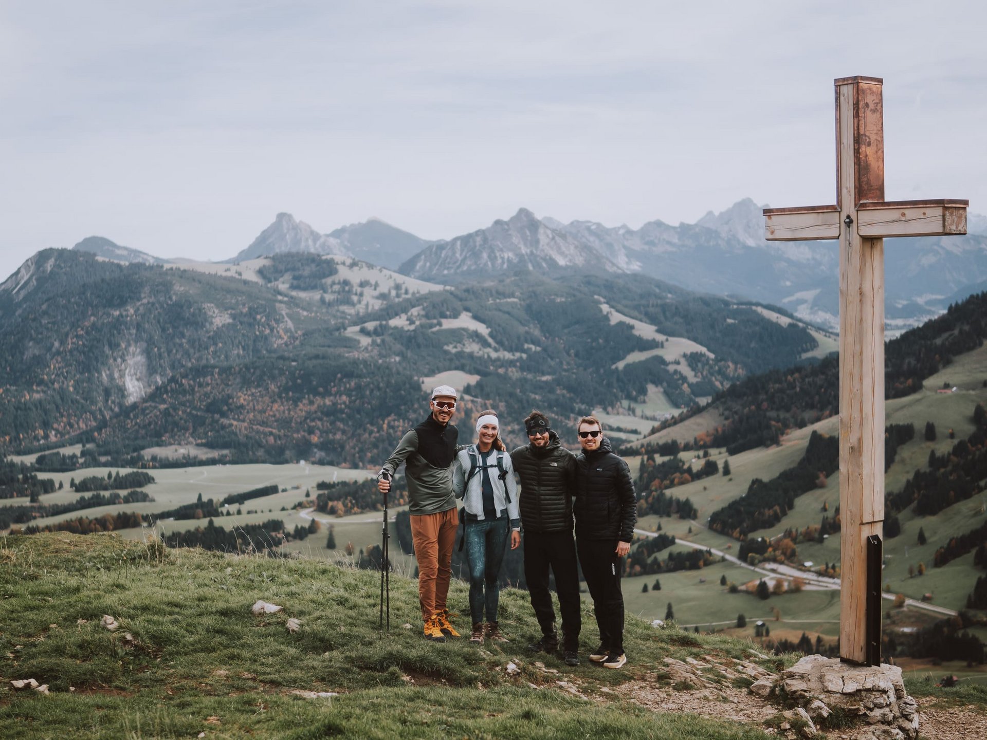 Alpin Lodges Oberjoch – Exklusive Ferienwohnungen mit Wellness Vier Wanderer stehen neben einem Gipfelkreuz mit Bergpanorama im Hintergrund